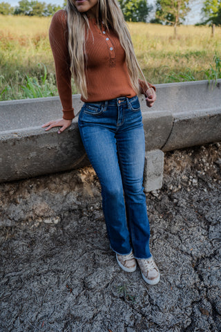 Person wearing an orange sweater and blue jeans standing by a concrete structure with a field in the background