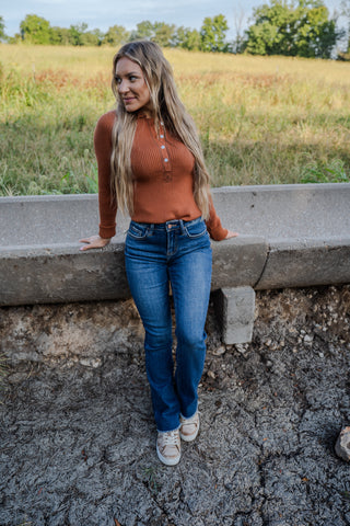 Woman in a rust-colored top and blue jeans standing on a concrete ledge with a field in the background