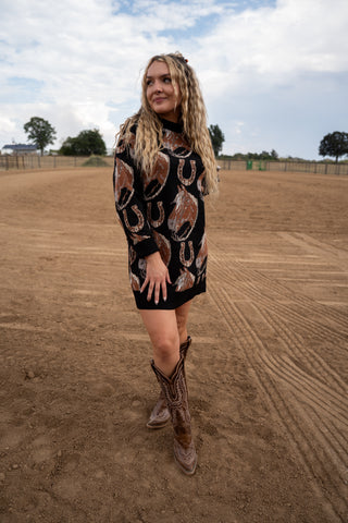 Woman wearing a patterned dress and cowboy boots standing on a dirt field with trees and a cloudy sky in the background.
