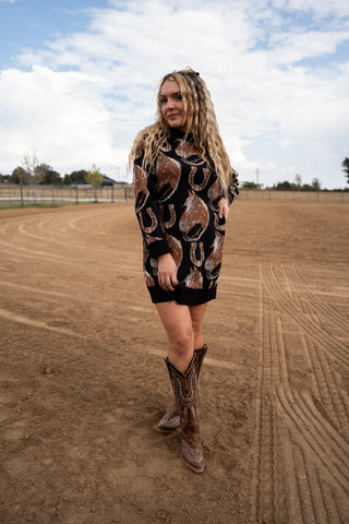 Woman wearing a patterned dress and cowboy boots standing on a dirt field with a cloudy sky.