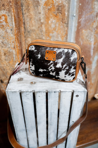 Cow print crossbody bag with brown leather accents on a white wooden crate against a rustic metal wall.