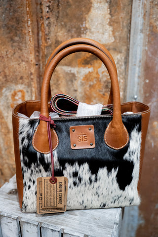 Handbag with cowhide pattern and brown leather handles against a rustic background