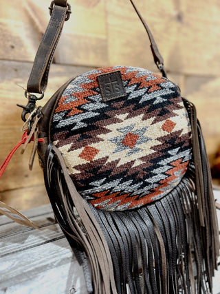 Round handbag with patterned top and fringed sides on a wooden surface