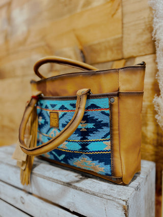 Brown leather handbag with a blue and green patterned flap on a wooden surface.