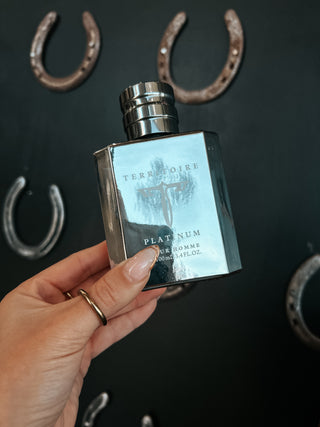 Hand holding a perfume bottle labeled 'Territorial Platinum' with horseshoe decorations in the background.