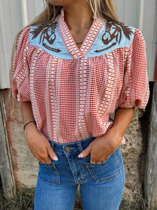 Person wearing a red and white checkered blouse with embroidered details and blue jeans, standing against a rustic wooden background.