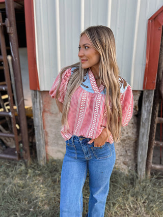 Woman wearing a red and white patterned blouse and blue jeans standing in front of a barn.