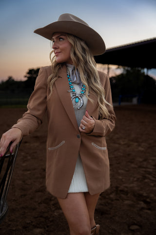 Woman in a cowboy hat and brown blazer standing on a dirt ground with a sunset sky.