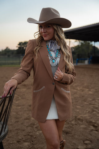 Woman in a cowboy hat and beige blazer standing in an outdoor setting.