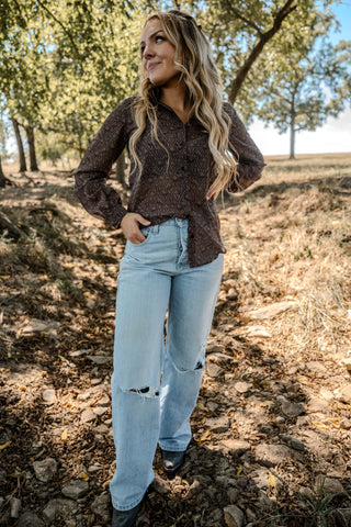 Woman standing outdoors in a natural setting wearing a brown top and light blue jeans.