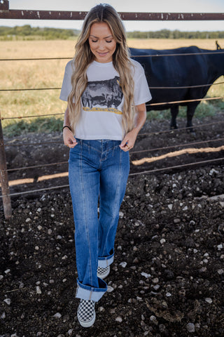 Woman standing in a field with a cow behind a fence, wearing a graphic t-shirt and jeans.