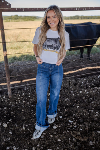 Woman standing in a field with a cow in the background, wearing a graphic t-shirt and jeans.