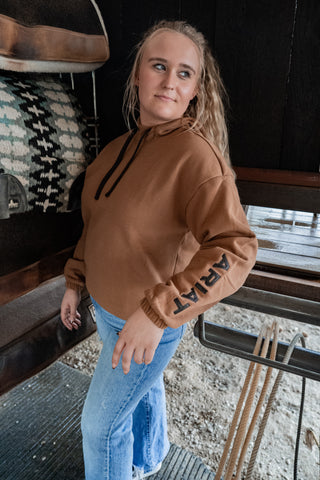 Woman wearing a brown sweatshirt with 'ART' on the sleeve, standing in a rustic setting.