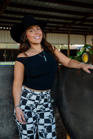 Woman wearing a black top and checkered pants with a cowboy hat, standing in a barn.
