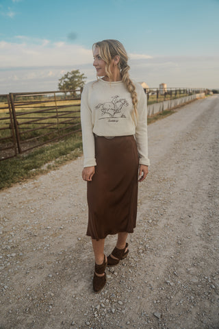 Woman standing on a dirt road wearing a white sweater with a graphic design and a brown skirt.