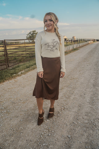 Woman standing on a dirt road wearing a cream sweater with a graphic design and a brown skirt.