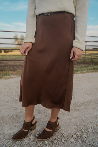 Person wearing a brown skirt and beige sweater standing on a gravel path with a fence and field in the background.