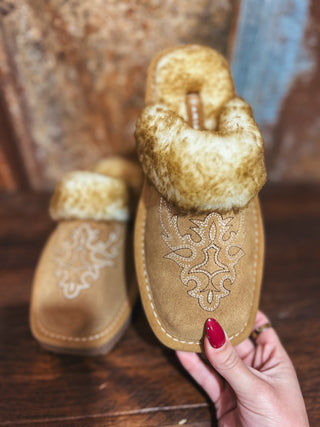 Pair of brown moccasin slippers with fur trim held by a hand on a wooden surface.