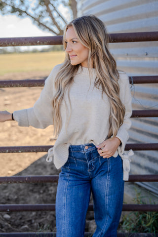 Woman wearing a beige sweater and blue jeans standing near a metal fence with a rural background.
