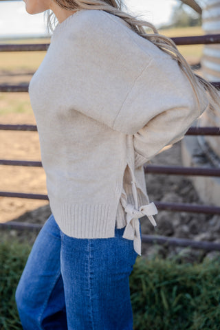 Person wearing a beige sweater with lace-up details and blue jeans, standing in front of a metal fence.