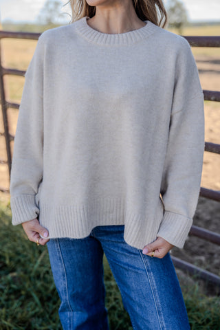 Person wearing a light gray sweater and blue jeans standing in front of a wooden fence.
