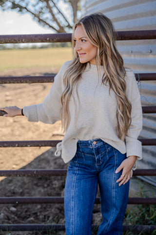 Woman in a beige sweater and blue jeans standing next to a metal fence with a rural background.