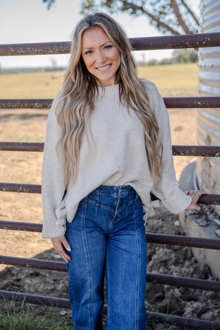 Woman wearing a beige sweater and blue jeans standing next to a metal fence with a rural background.