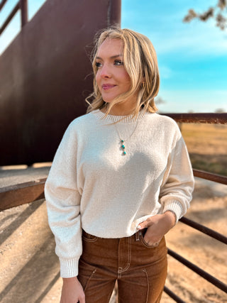 Woman wearing a white sweater and brown pants standing outdoors with a scenic background.