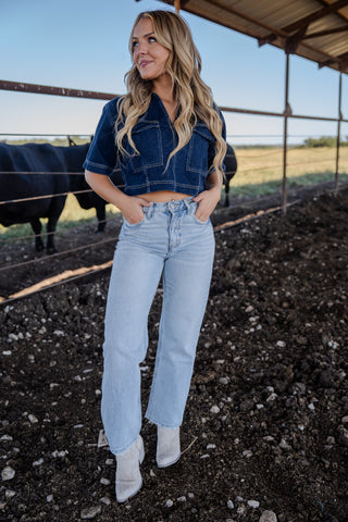Woman standing in a barn wearing a denim crop top and light blue jeans.