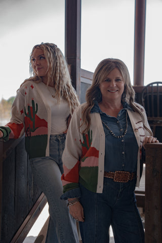 Two women standing indoors, one wearing a colorful cardigan with cactus design.