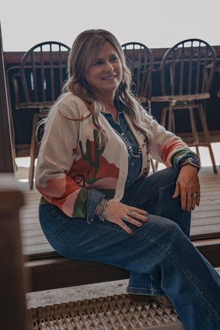 Woman sitting on a wooden bench wearing a colorful cardigan with a cactus design.