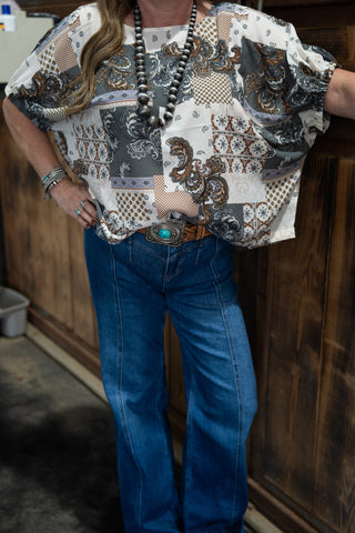 Person wearing a patterned shirt and blue jeans leaning against a wooden wall.