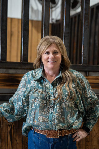 Woman wearing a patterned shirt and blue jeans standing in front of wooden beams.