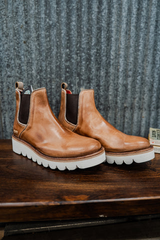 Pair of brown leather boots with white soles on a wooden surface against a textured gray wall.
