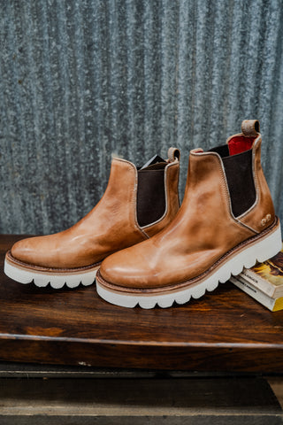 Pair of brown leather boots with white soles on a wooden surface against a textured gray wall.