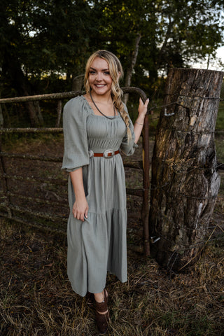 Woman in a long light gray dress standing next to a wooden fence with trees in the background