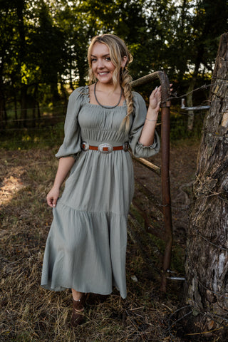 Woman in a green dress standing outdoors with a hoe, surrounded by trees and natural light.