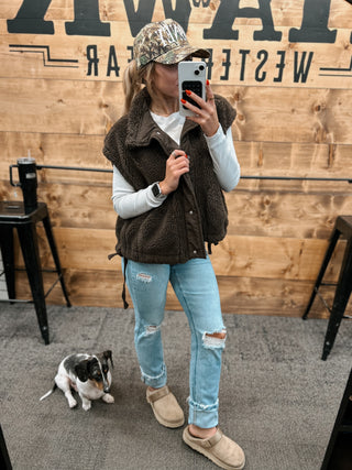 Person taking a mirror selfie wearing a brown vest, white shirt, blue jeans, and beige boots with a small dog on the floor.