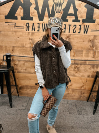 Person taking a mirror selfie wearing a brown vest, light blue jeans, and a patterned hat in front of a wooden wall with 'TAMW' branding.