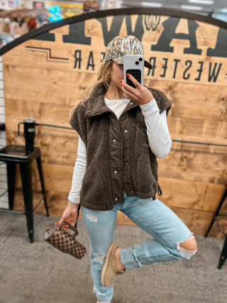 Person wearing a brown vest, light blue jeans, and a patterned cap in front of a wooden wall with text.