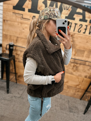 Person wearing a brown fleece vest and camouflage cap taking a selfie.