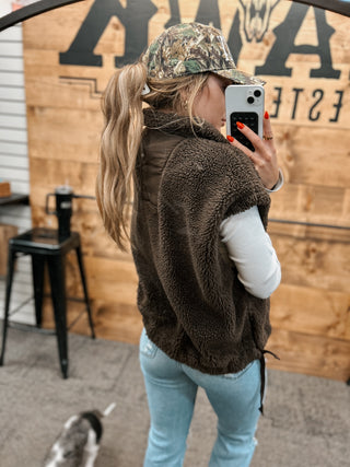 Person wearing a brown fleece jacket and camouflage hat taking a selfie in front of a wooden wall with text.
