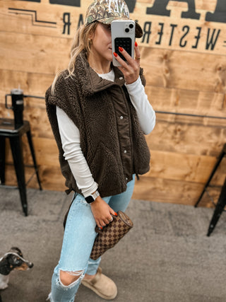 Person wearing a brown fleece vest, white long-sleeve shirt, light blue jeans, and camouflage cap, holding a phone and a patterned bag.