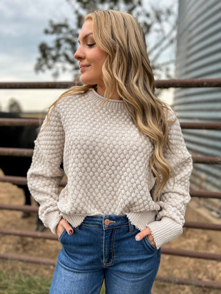 Woman wearing a textured beige sweater and blue jeans standing outdoors.