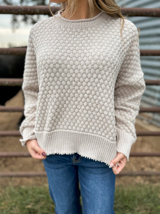 Person wearing a textured beige sweater and blue jeans standing in front of a metal fence.