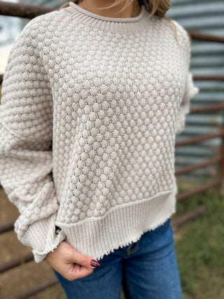 Person wearing a textured beige sweater with blue jeans against a rustic metal fence background