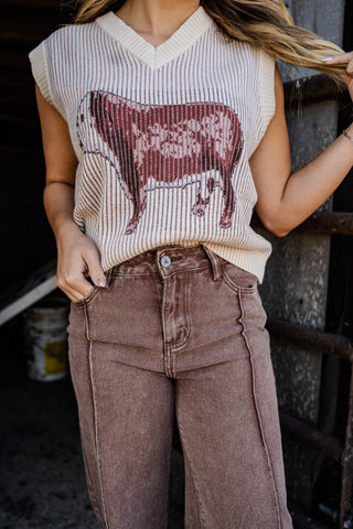 Person wearing a sleeveless top with a cow print and high-waisted brown pants in an indoor setting.