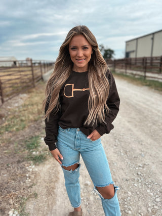 Woman wearing a black sweatshirt with a logo and light blue jeans standing on a dirt road.
