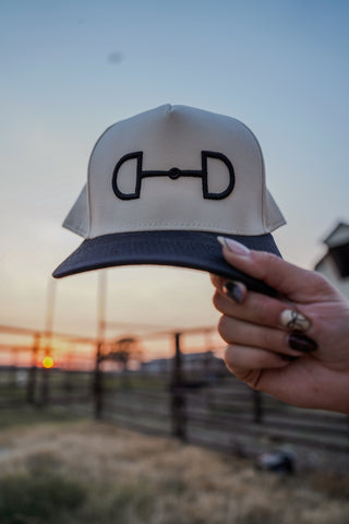 White cap with black logo held by a hand against a sunset background