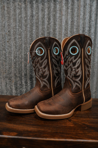 Pair of brown cowboy boots with decorative patterns on a wooden surface and metal wall background